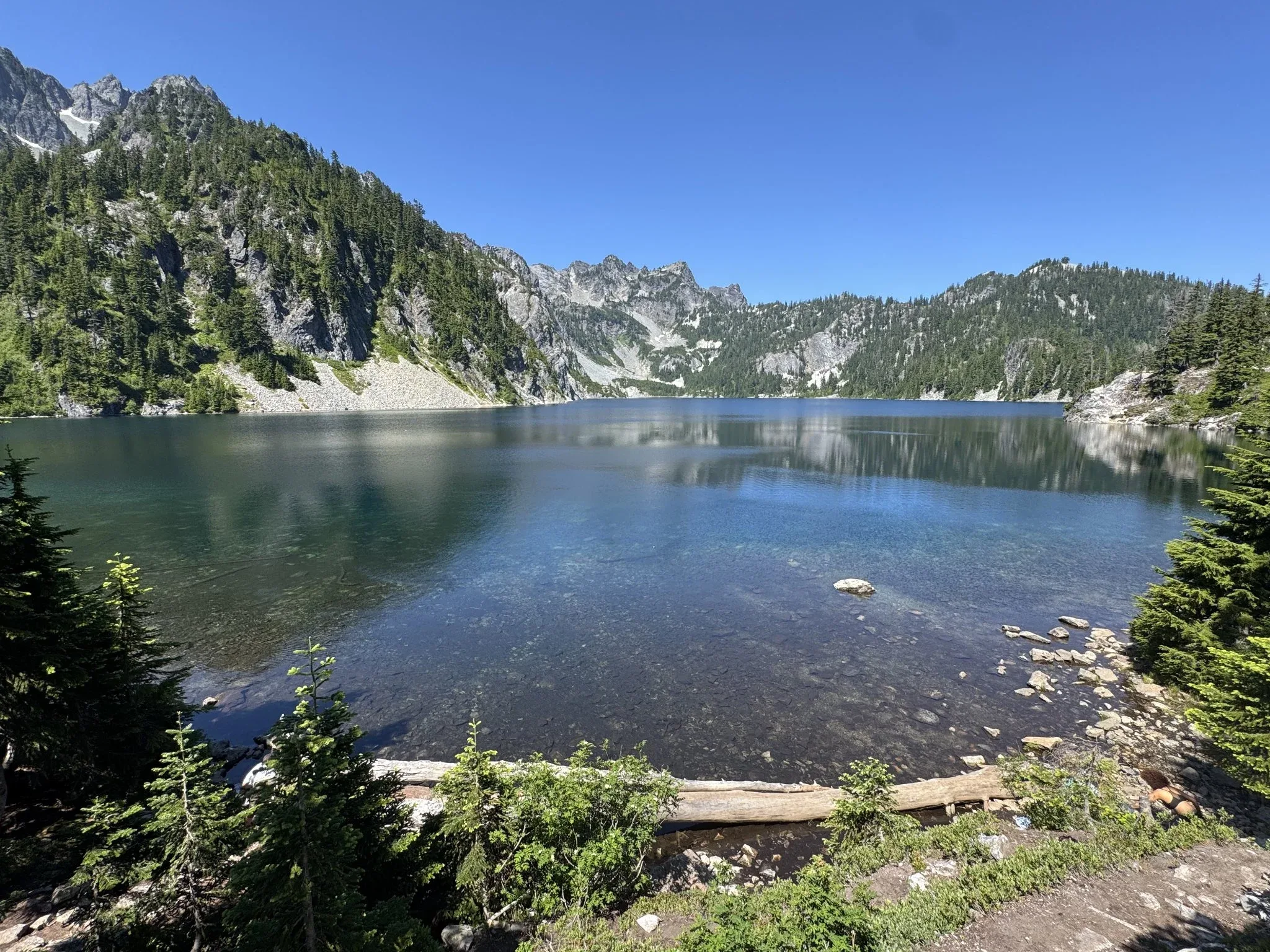 The shore of Snow Lake with shallow water showing the rocks and dirt below the water. A mountainous ridge is around the back end of the lake.